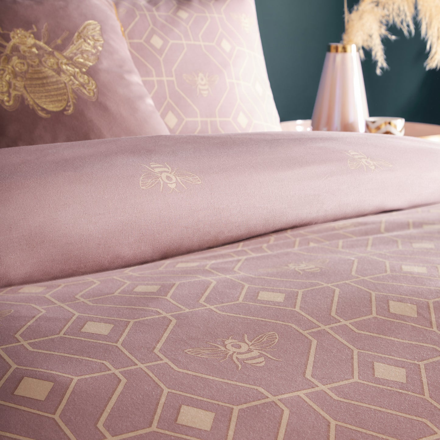 Close up of beautiful luxury duvet cover bedding set with a geometric pattern and small bees in each geometric shape, side table with pink vase and natural flora, teacup. Top of duvet cover is folded over to show reversible side. Bedding is in a blush pink colour.