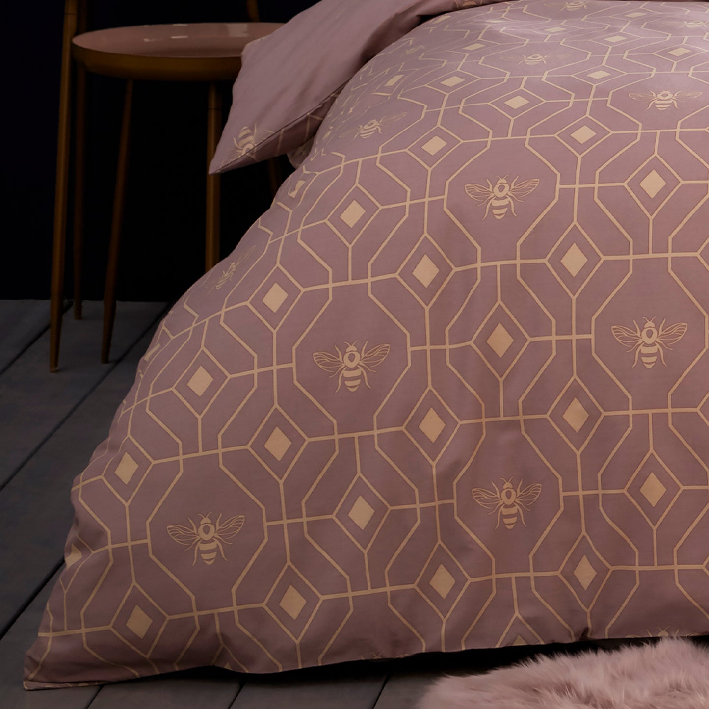 Close up of corner of a bed showing a beautiful luxury duvet cover bedding set with a geometric pattern and small bees in each geometric shape, small wooden bed side table. Bedding is in a blush pink colour.