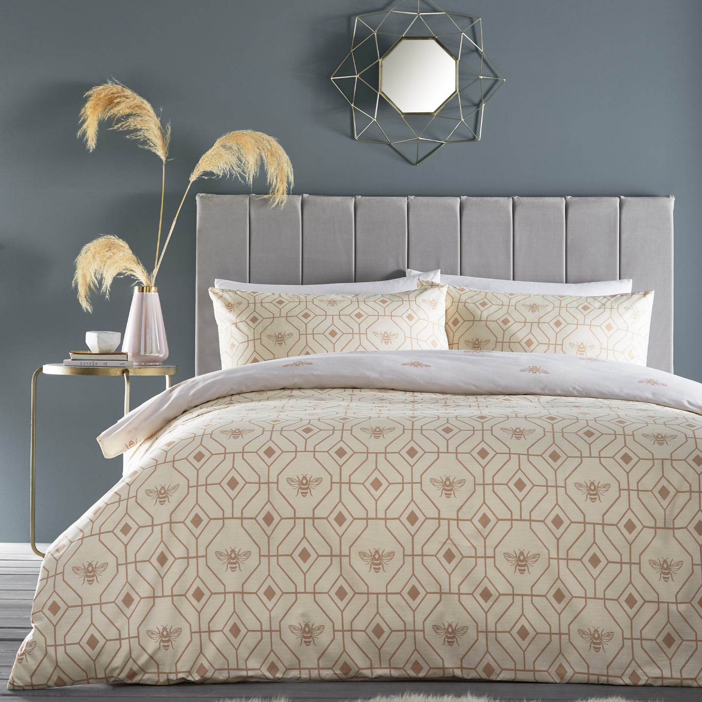 Beautiful luxury duvet cover bedding set with a geometric pattern and small bees in each geometric shape, grey headboard and sliver accent mirror above on the wall. Bedding is in a colour. champagne.