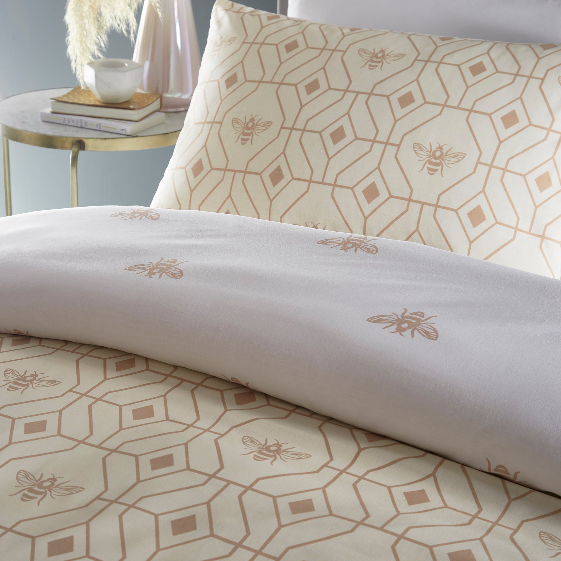 Close up of a beautiful luxury duvet cover bedding set with a geometric pattern and small bees in each geometric shape, grey headboard  and side table. Duvet is folded over to show white reversible side . Bedding is in a champagne colour.