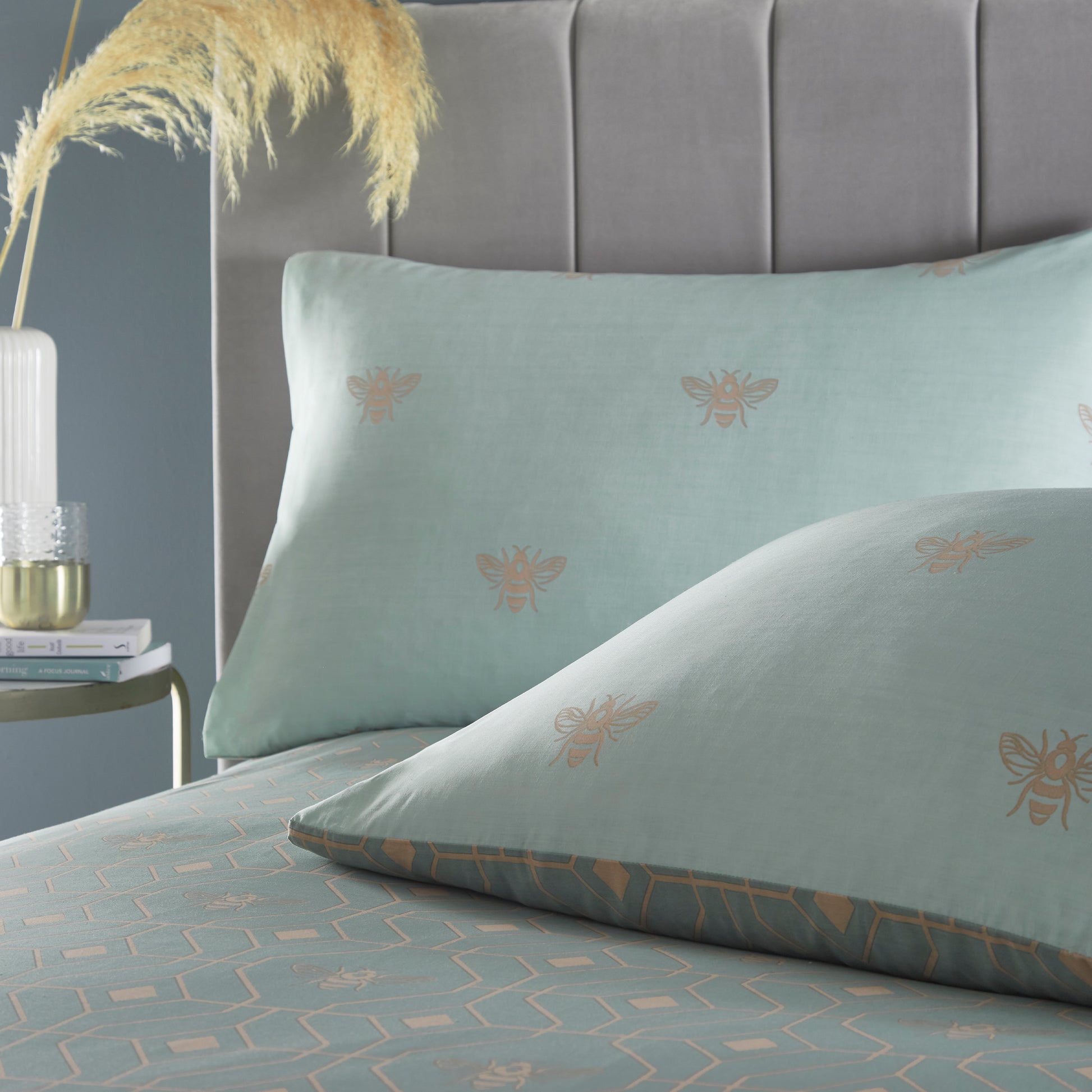 Pillow close up of beautiful luxury duvet cover bedding set with a geometric pattern and small bees in each geometric shape, grey headboard and natural flora in a vase on the side stand.  Bedding is in eau de nil sage colour. 