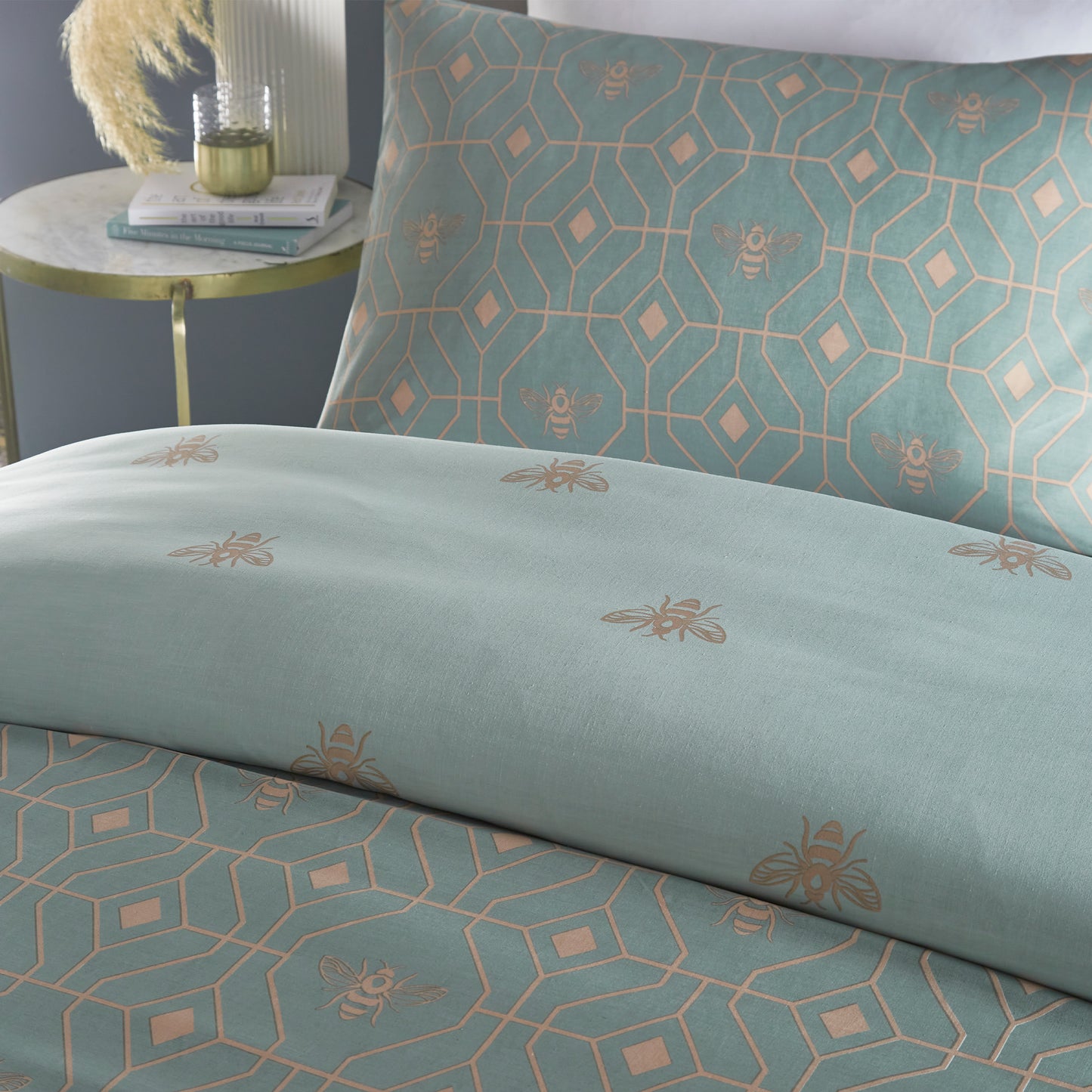 Close up of beautiful luxury duvet cover bedding set with a geometric pattern and small bees in each geometric shape, grey headboard and bedside table with two books and a tumbler. Bedding is in eau de nil sage colour. 