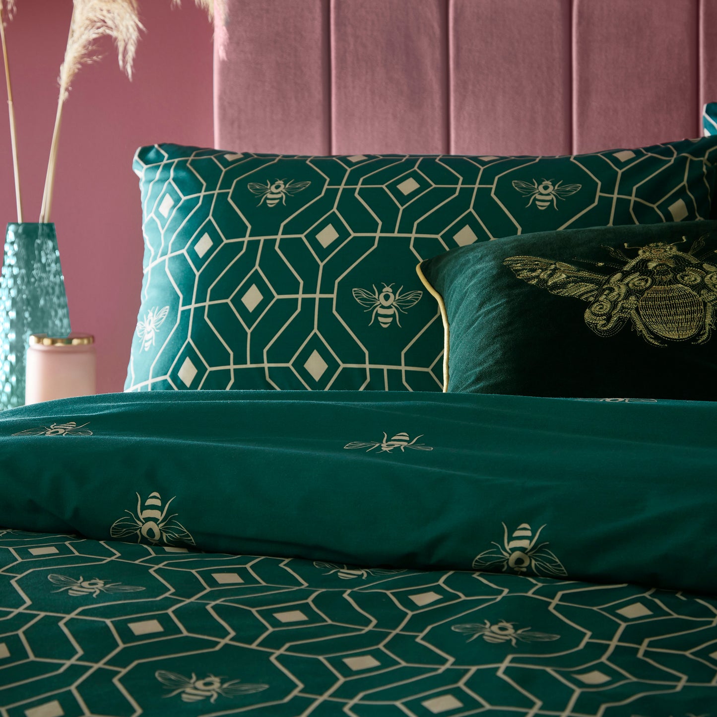 Close up of two pillows in a beautiful luxury duvet cover bedding set with a geometric pattern and small bees in each geometric shape, burgundy headboard and side table with natural flora displayed. Bedding is in a dark emerald green colour.