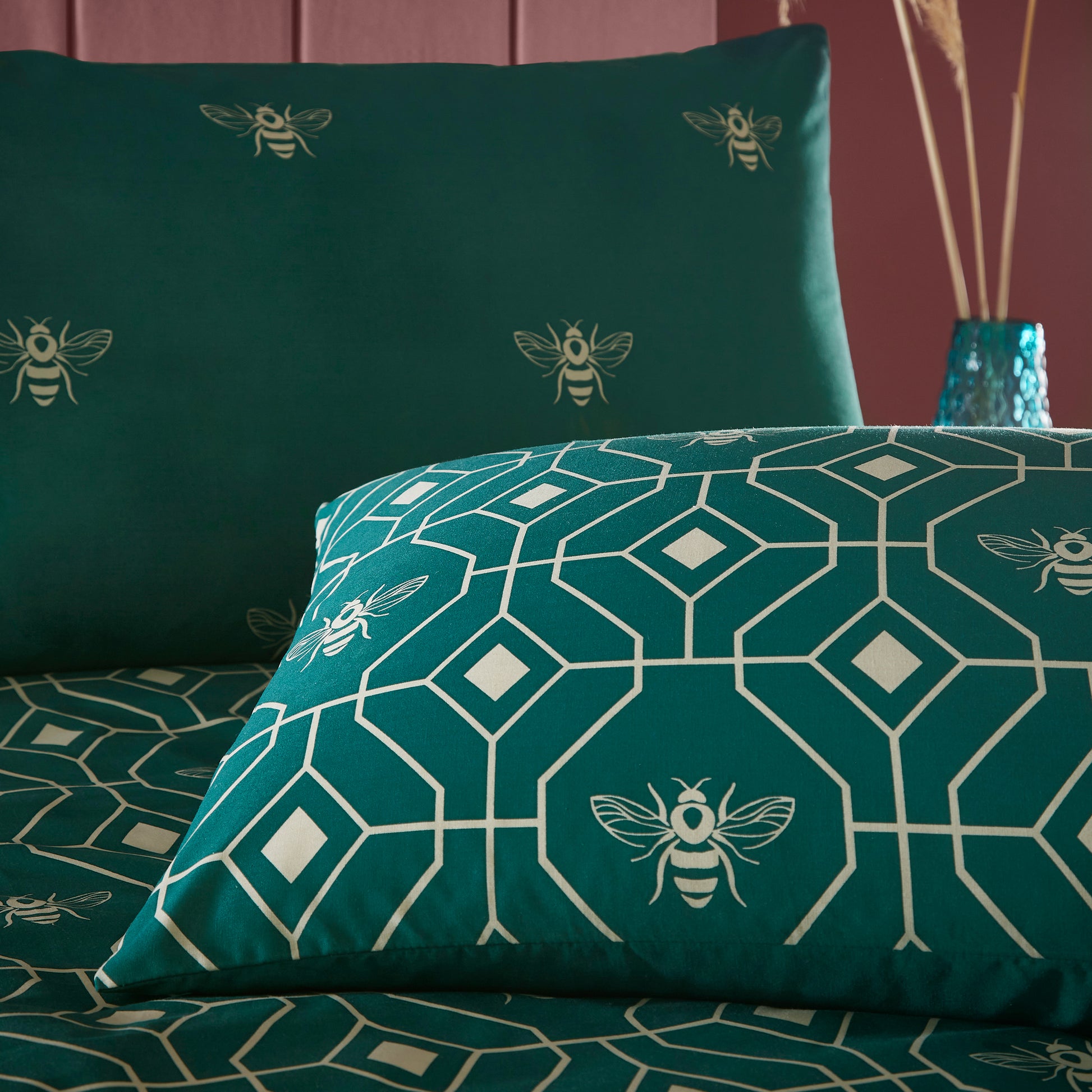 Close up of two pillows in a beautiful luxury duvet cover bedding set with a geometric pattern and small bees in each geometric shape, burgundy headboard. Bedding is in dark green emerald colour.