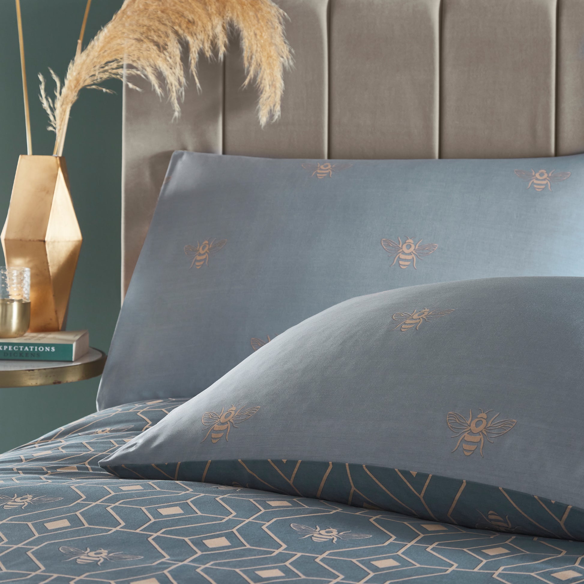 Close up of two beautiful luxury duvet cover bedding set with a geometric pattern and small bees in each geometric shape, with a gold trim, grey headboard and night stand with vase, book and a glass. Bedding is in a French Blue colour. Pillows show reversible side.