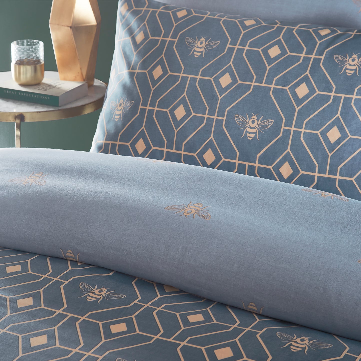 Close up of beautiful luxury duvet cover bedding set with a geometric pattern and small bees in each geometric shape, with a gold trim, grey headboard, night stand with a vase, book, and a glass. Bedding is in a French Blue colour.