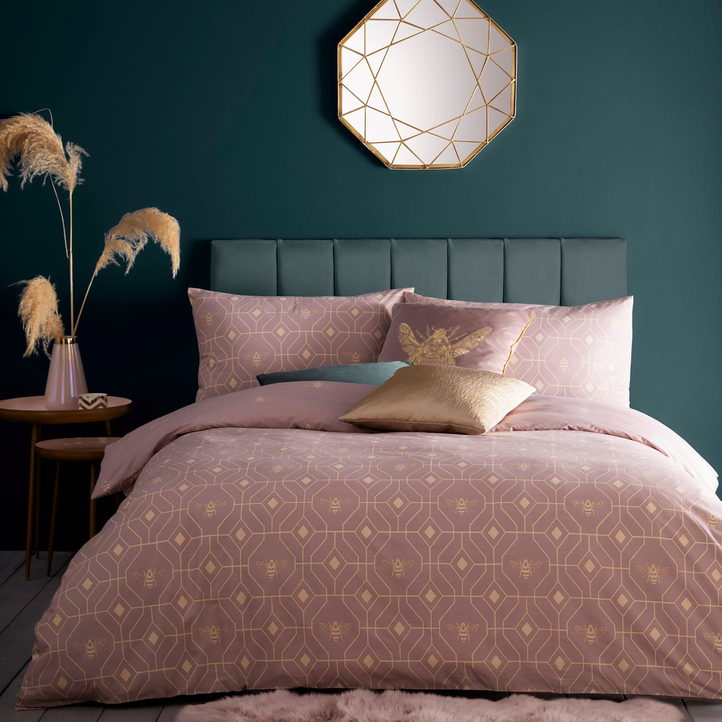 Beautiful luxury duvet cover bedding set with a geometric pattern and small bees in each geometric shape, green headboard and gold asymmetric accent mirror above on the wall. Bedding is in a blush pink colour. Side table with natural flora in a pink vase and a teacup and scatter cushion.