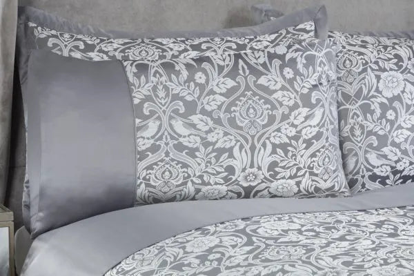 Close up of two pillows on a double bed with grey headboard from a side view showing one half of the bed  displaying the Harrow Collection Embellished Jacquard Duvet Double Bedding Cover Set in silver with a white sheen embroidery in a floral pattern. 