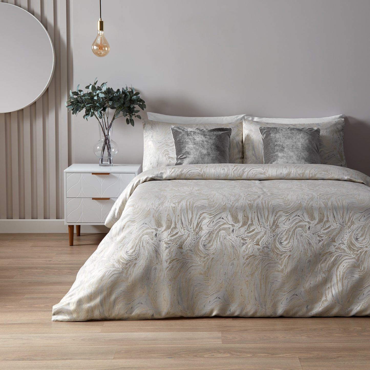 Double bed displaying a luxury bedding set with an intricate marbled pattern, featuring shades of oyster, beige, and blue. Accent cushions in a deep grey velvet style. Short white wooden side table with clear vase and green foliage. Hanging tinted light bulb from the ceiling and large round mirror on the wall to the side. 