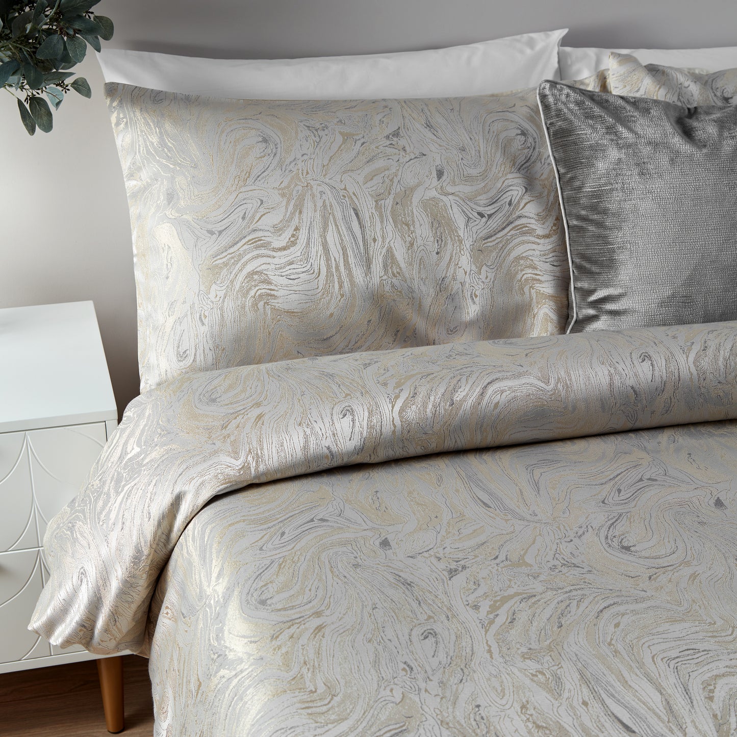 Close up of one side of the double bed displaying a luxury bedding set with an intricate marbled pattern, featuring shades of oyster, beige, and blue. One accent cushion in a deep grey velvet style displayed. Short white wooden side table with clear vase and green foliage can be half seen to one side. 