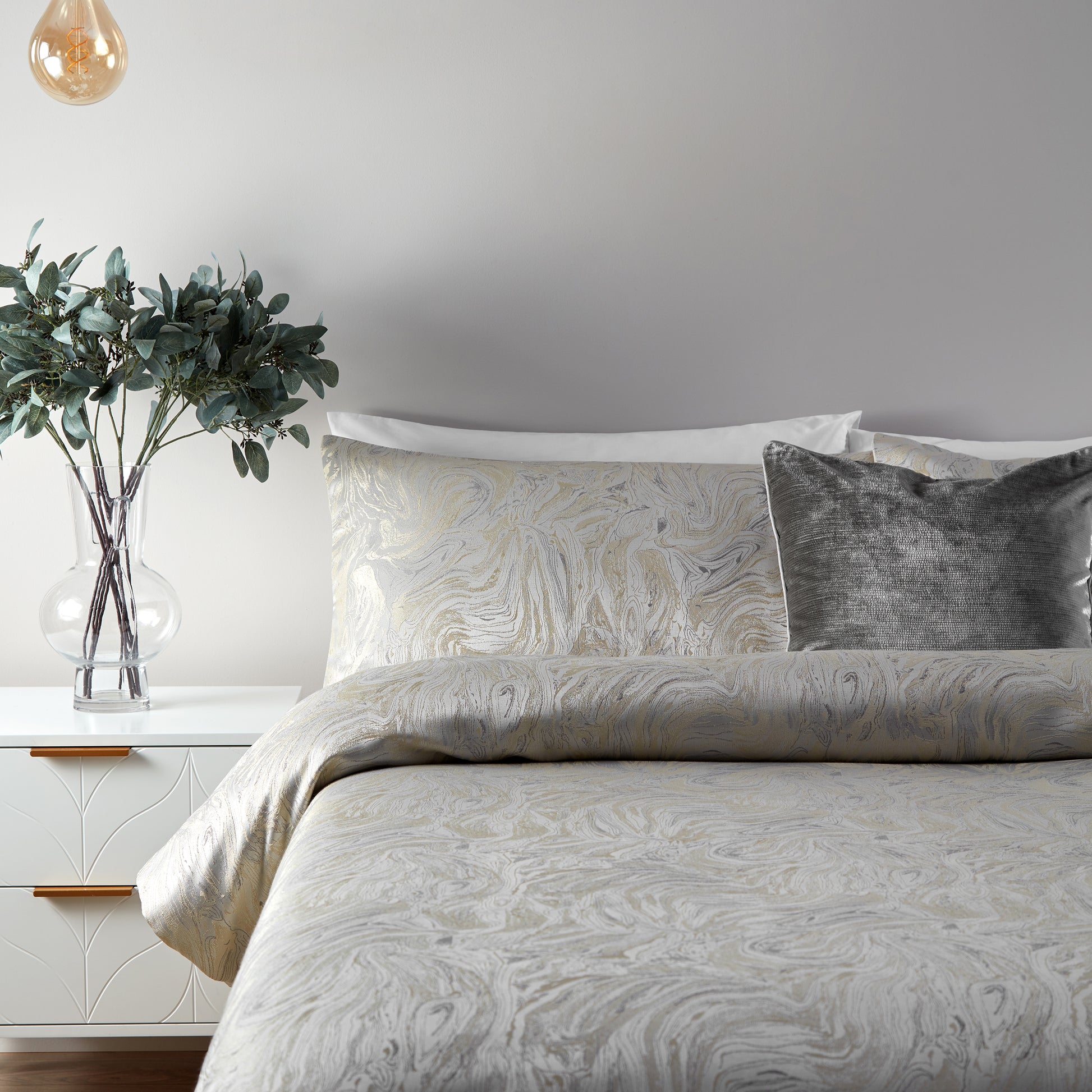 Close up of one side and centre of a double bed displaying a luxury bedding set with an intricate marbled pattern, featuring shades of oyster, beige, and blue. Accent cushion in a deep grey velvet style in the centre. Short white wooden side table with clear vase and green foliage stems visible to one side. Hanging tinted light bulb from the ceiling above the foliage.