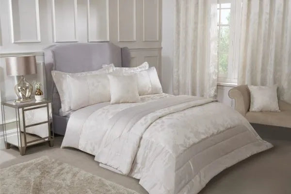 Double bed with grey headboard from a side view in front of a window displaying the Meadows Collection Embellished Jacquard Duvet Double Bedding Cover Set in Cream. Chaise lounge in a neutral tone with accent cushion against the wall. Mirrored side table with two drawers and large lamp with clear lamp and neutral shade. Matching bedspread also displayed on the end of the bed.