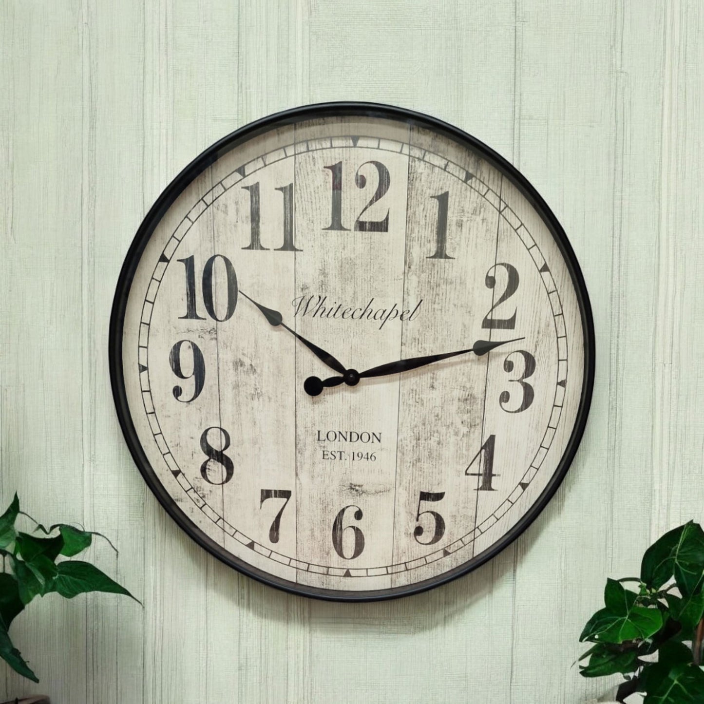 Round antique style wall clock with a faint sage striped natural wallpaper background and green foliage visible in each bottom corner