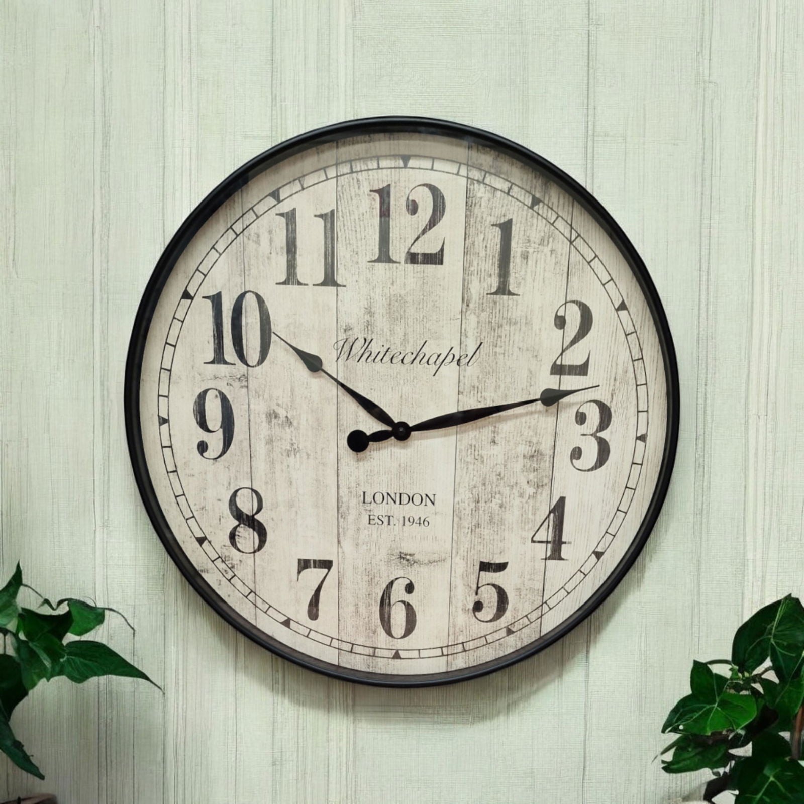 Round antique style wall clock with a faint sage striped natural wallpaper background and green foliage visible in each bottom corner