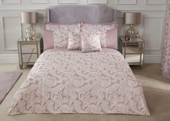 Double bed with grey headboard from a central view displaying the Duchess Collection Embellished Jacquard Duvet Double Bedding Cover Set in Blush Pink. Two  mirrored side table each side with two drawers and a large lamp with clear lamp and neutral shade. Accent cushions displayed in same pattern.